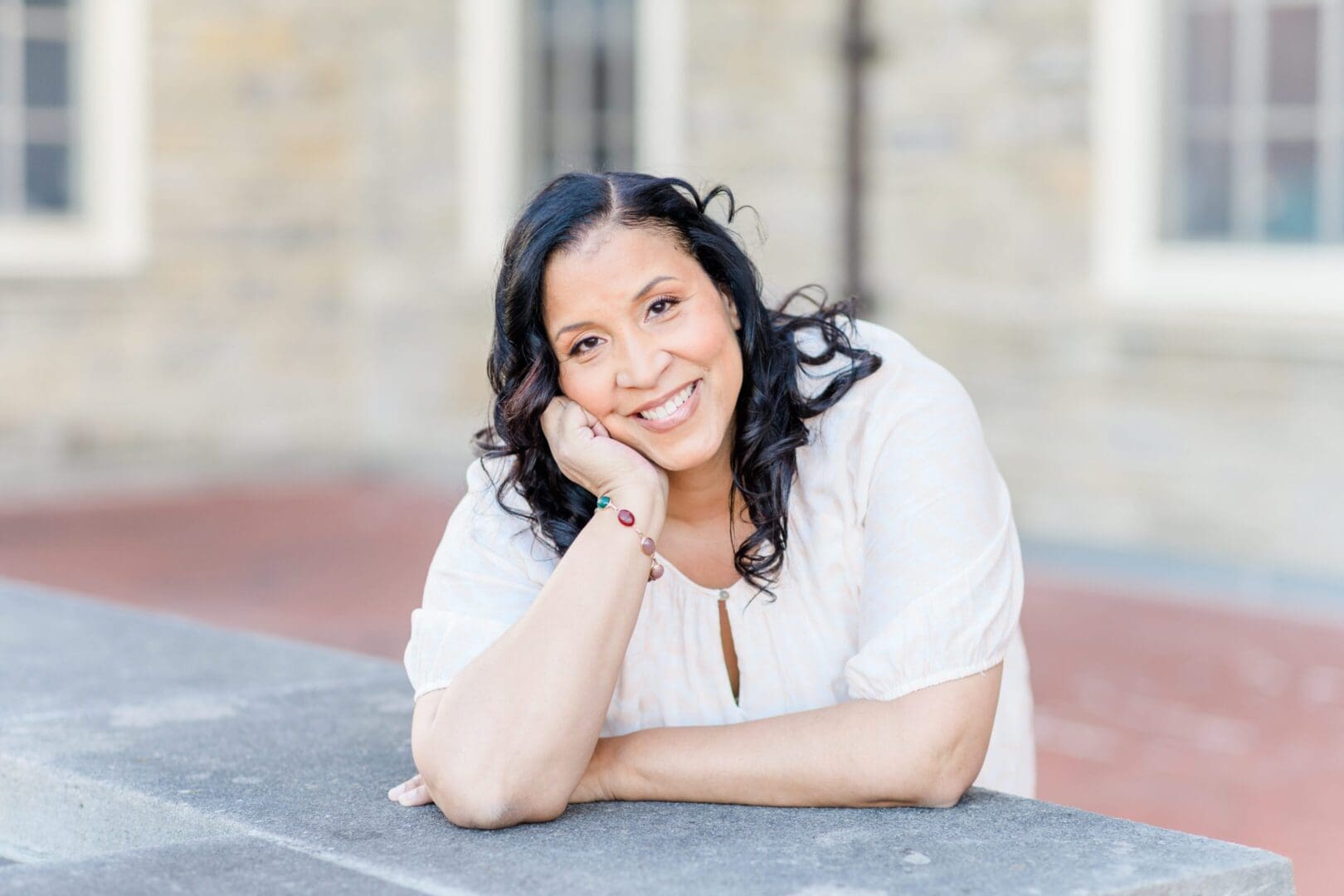 Smiling woman with long dark hair leaning on a stone surface outdoors.